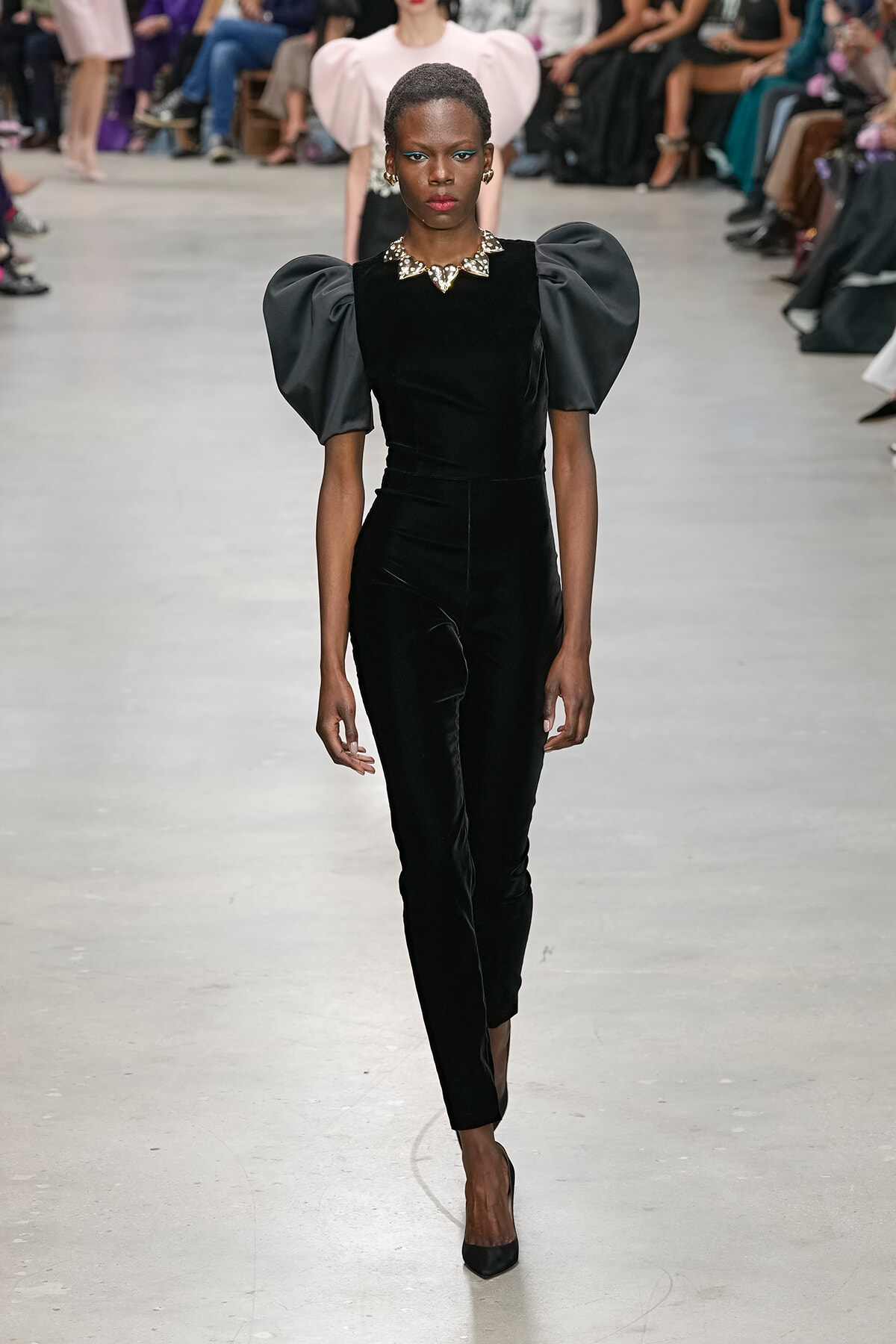 Model walks a fashion runway wearing a black velvet jumpsuit with dramatic puff sleeves and a jeweled collar necklace; audience blurred in background.