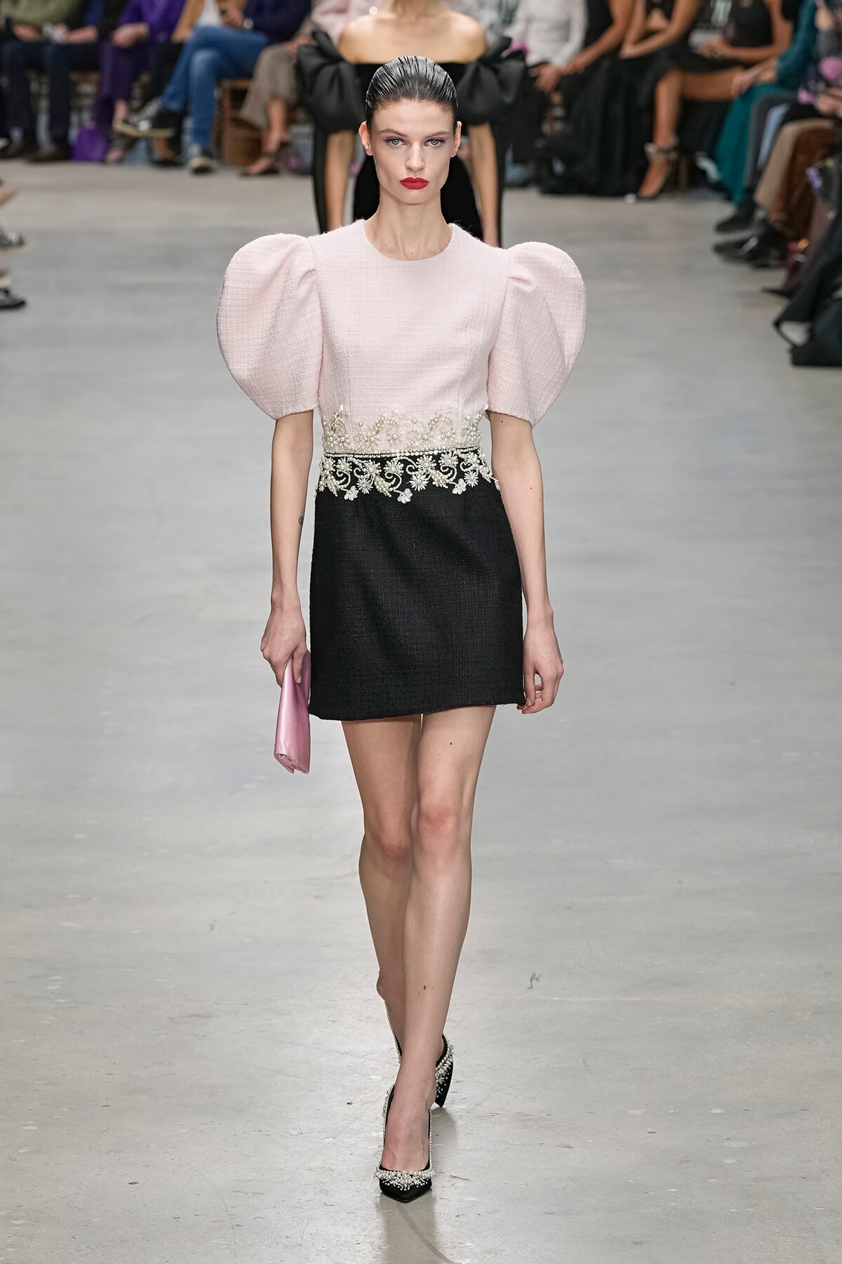 Model walks a fashion show wearing a pale pink top with dramatic puff sleeves and a black skirt with an ornate lace belt, holding a pink clutch.