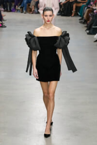 Model walks the runway in a black off-the-shoulder mini dress with oversized bow sleeves.
