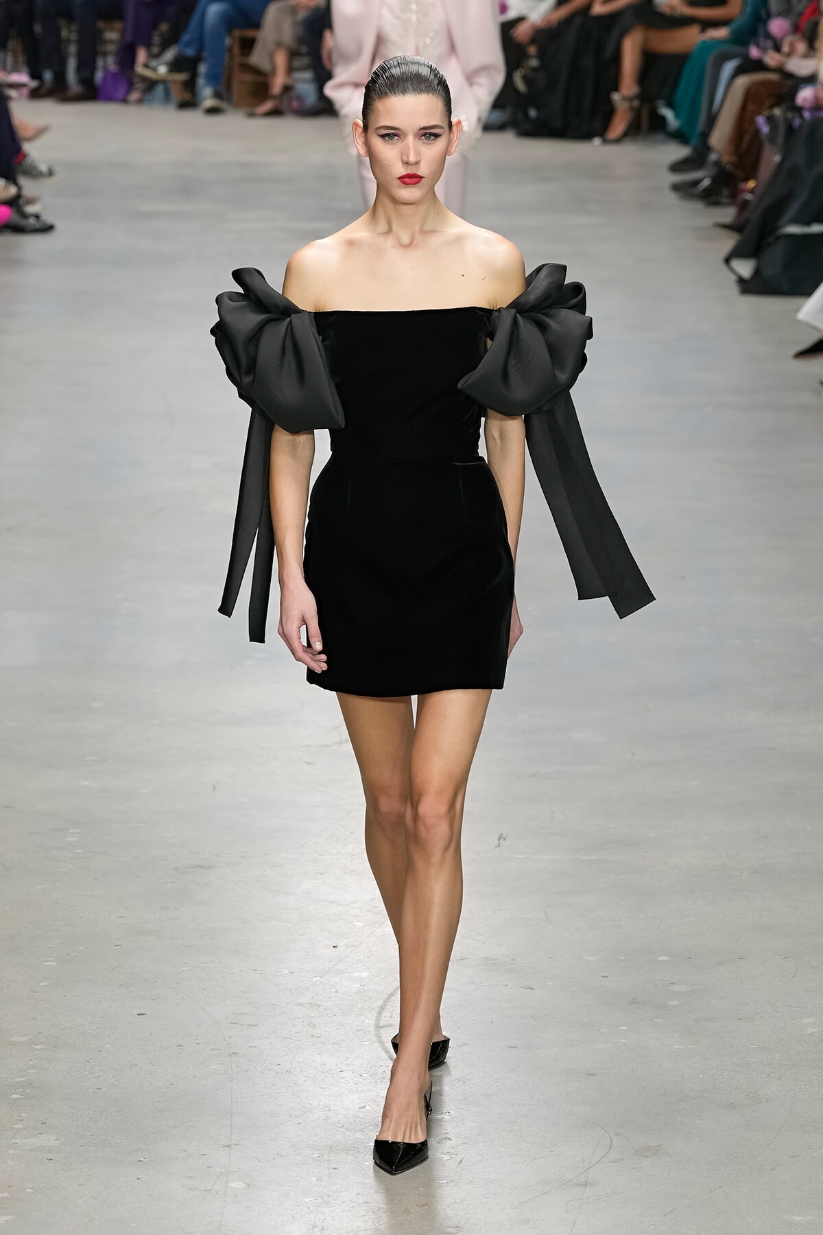 Model walks the runway in a black off-the-shoulder mini dress with oversized bow sleeves.