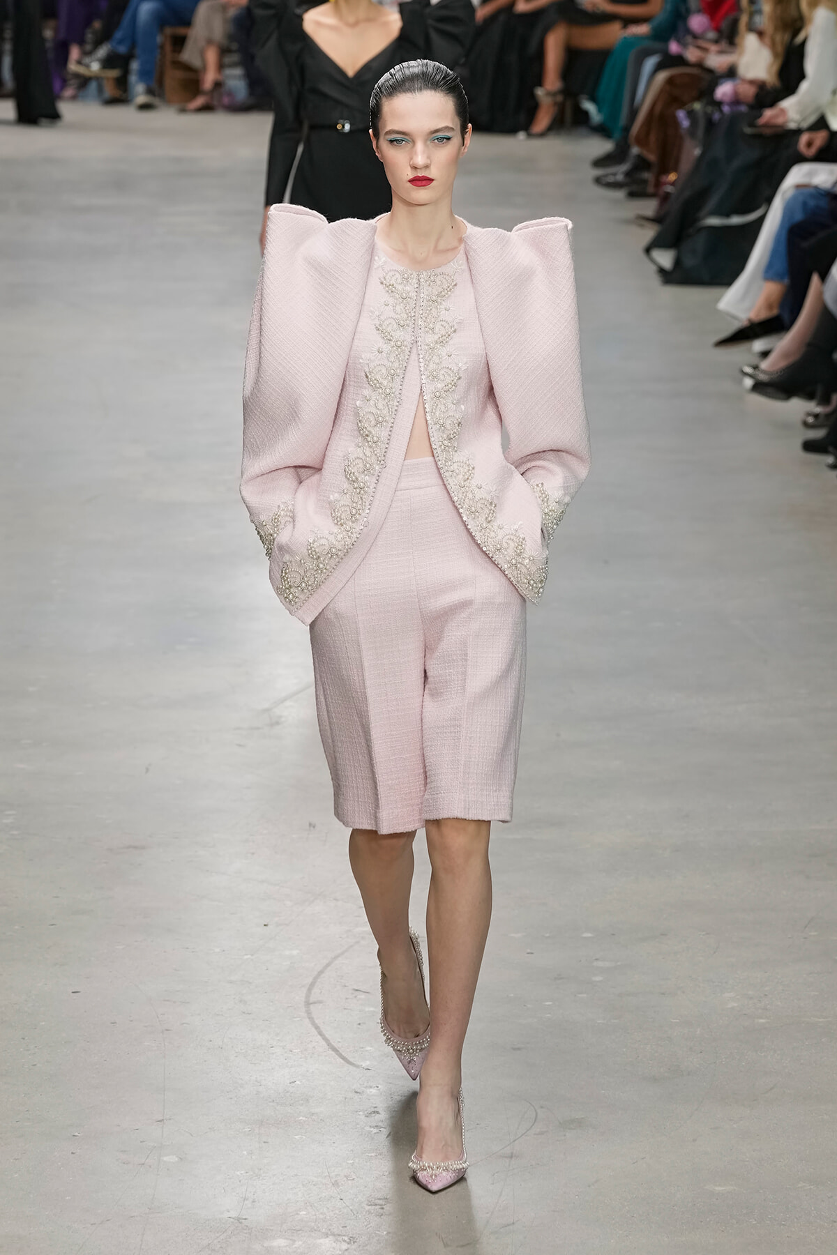 Model walks the runway in a pale pink, textured suit with oversized shoulders and ornate embroidery along the front and cuffs, paired with pink heels.