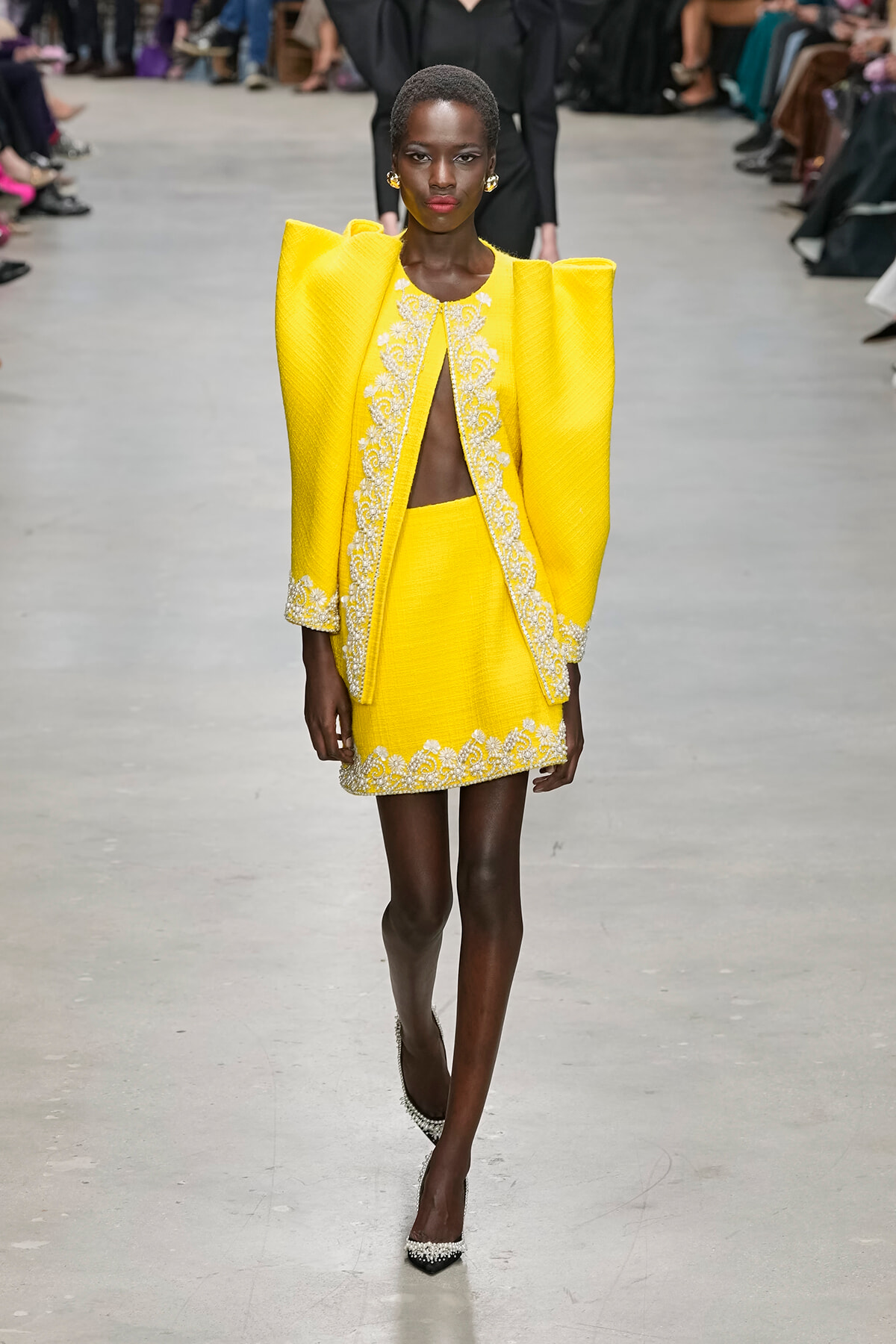 Model walks a runway in a bright yellow outfit with sculpted shoulders and white embroidered trim from neck to hem on a mini dress and jacket edges.