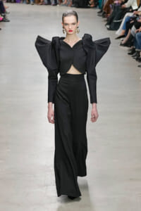 Model walks a runway in a black high-fashion outfit with dramatic oversized puff sleeves and cropped top, gold earrings, red lips.