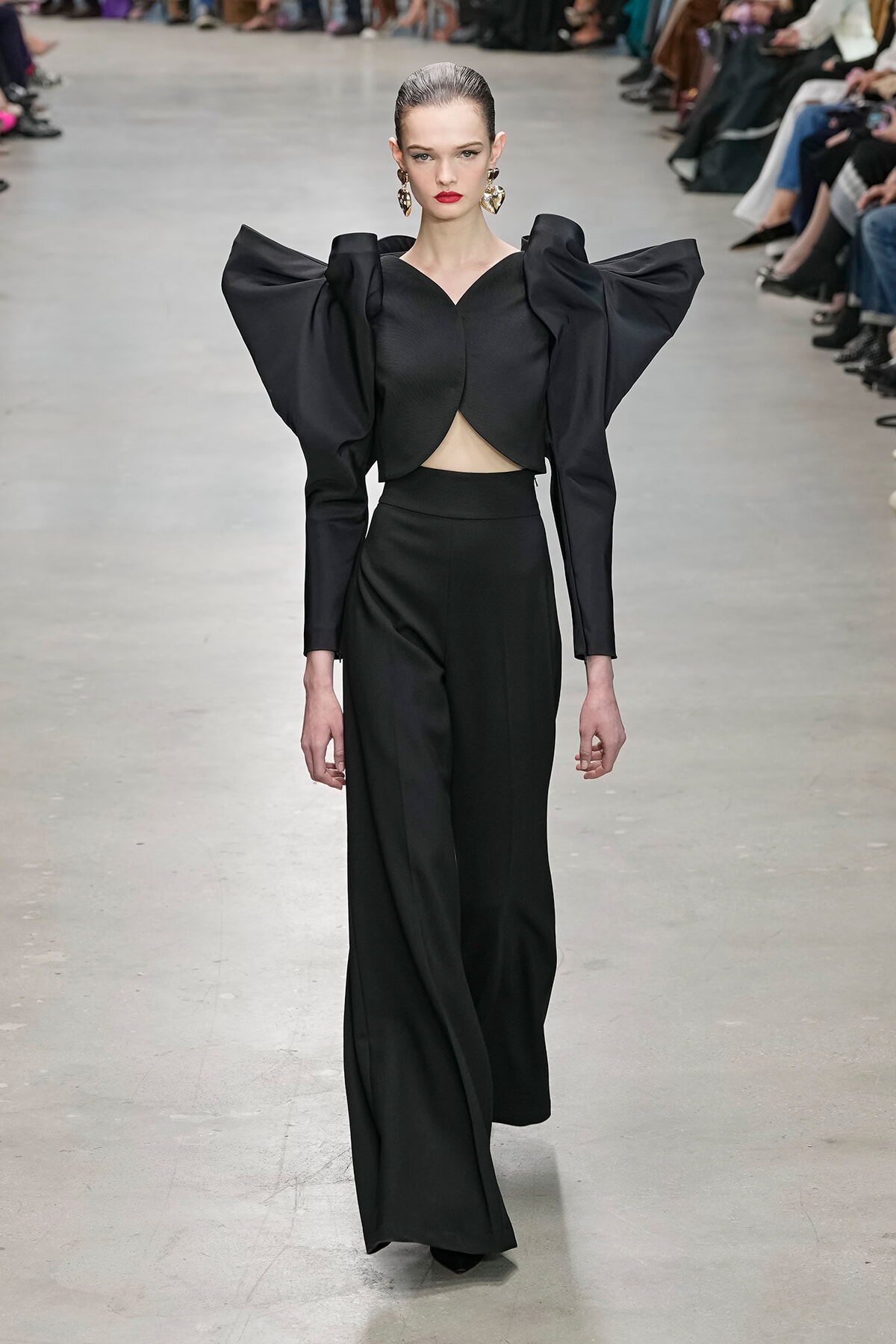 Model walks a runway in a black high-fashion outfit with dramatic oversized puff sleeves and cropped top, gold earrings, red lips.