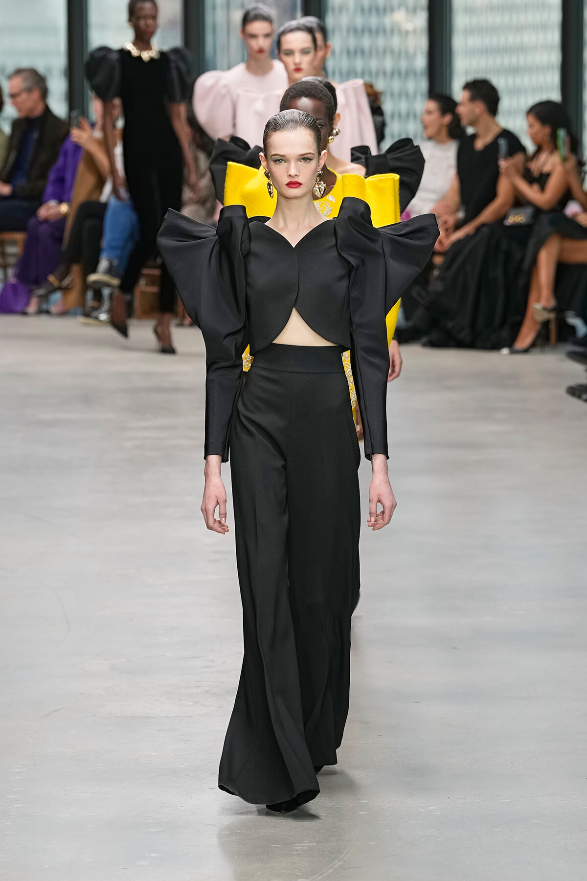 Model on a runway wearing a black jumpsuit with oversized sculptural sleeves and a midriff cutout; audience seated in background.