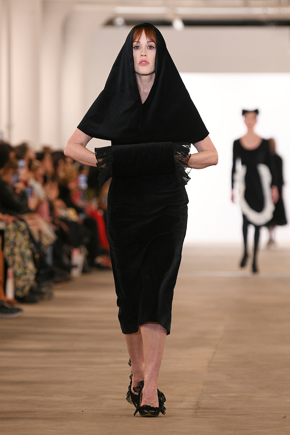 Runway model in an all-black ensemble with a hooded cape and lace-trim sleeves walking down the catwalk.