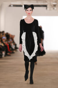 Model walking the runway in a black velvet dress with white lace ruffle panels and a large black bow headband.