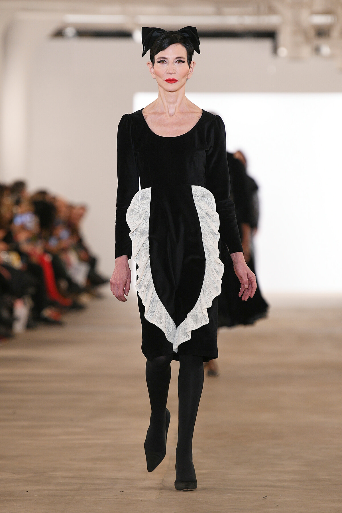 Model walking the runway in a black velvet dress with white lace ruffle panels and a large black bow headband.