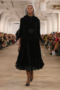 Model walks the runway in a long black velvet dress with a ruffled collar and belt, audience in the background.