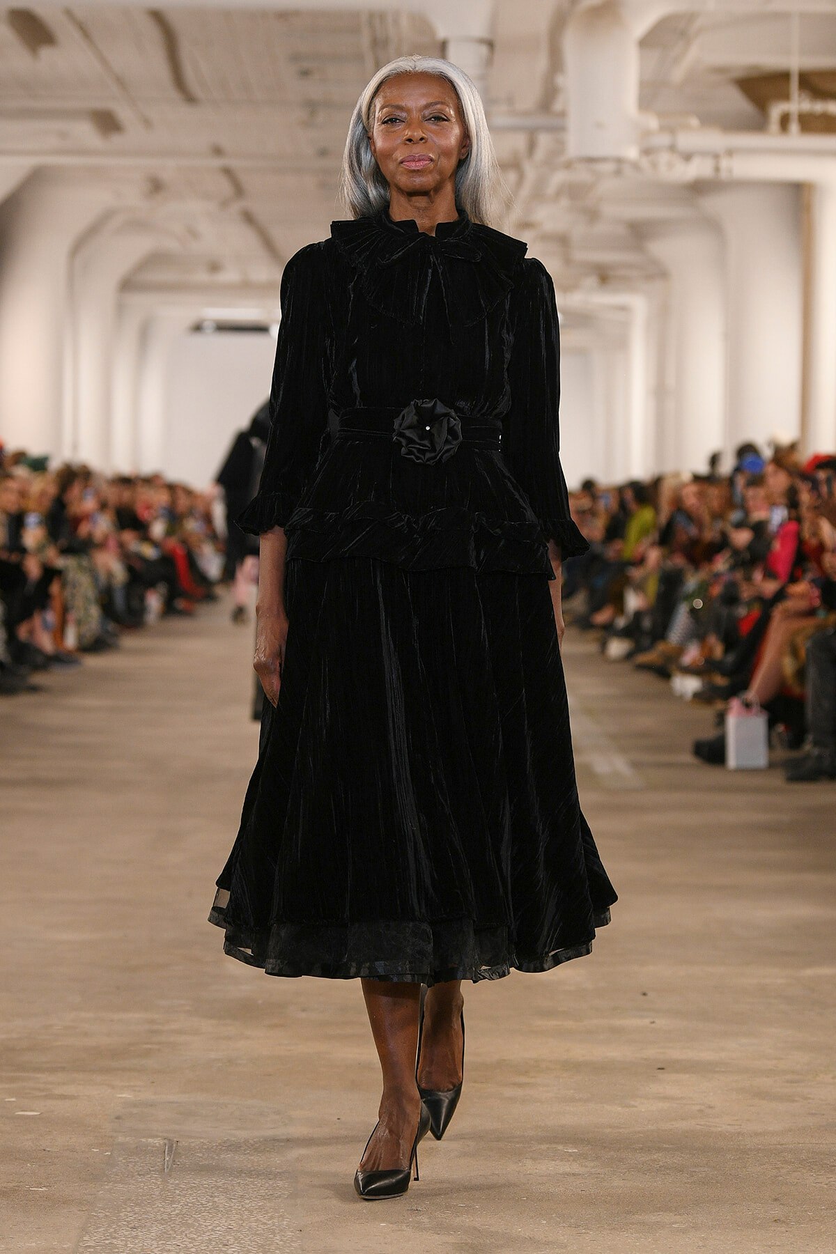 Model walks the runway in a long black velvet dress with a ruffled collar and belt, audience in the background.