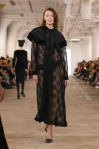 Model walking a runway in a sheer black lace floor-length dress with a cape-like overlay, surrounded by seated spectators.