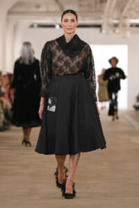 Model walking the runway in a sheer black lace blouse with a high neckline and a dark A-line skirt with a patch detail on the hip.