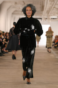 Model walks the runway in a black velvet outfit with white floral embroidery and matching pants, during a fashion show.