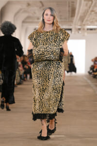 Model walking a fashion show in a full-length leopard-print faux fur dress with black lace trim and ruffled hem, hands in pockets pose.