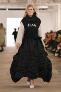 Model walking a fashion runway in a voluminous black ballgown with beige knit sleeves and a high neck; the sweater top reads 'HAG'.