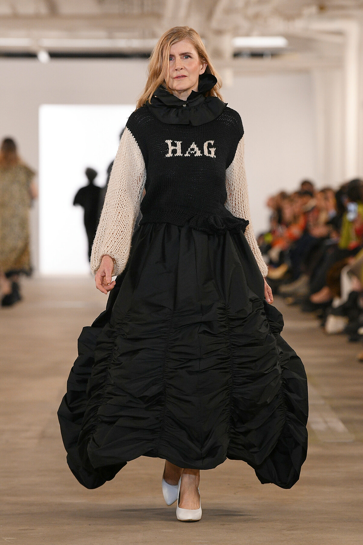 Model walking a fashion runway in a voluminous black ballgown with beige knit sleeves and a high neck; the sweater top reads 'HAG'.