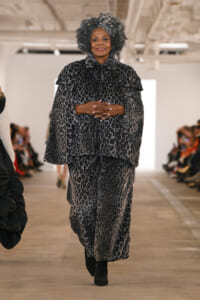 Model walking a runway in a gray leopard-print fur coat and matching skirt, hands clasped in front, confident expression.