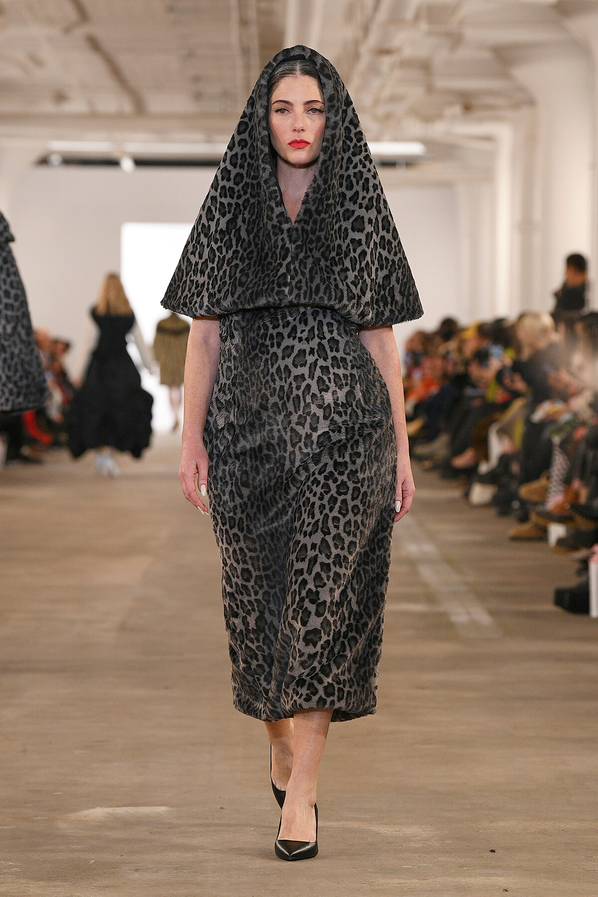 Model walking down a fashion runway in a gray leopard-print hooded cape over a matching dress, paired with black heels.