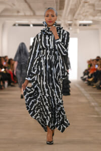 Model walks the runway in a black-and-white striped, ruffled dress with a high collar and belt; audience blurred in the background.