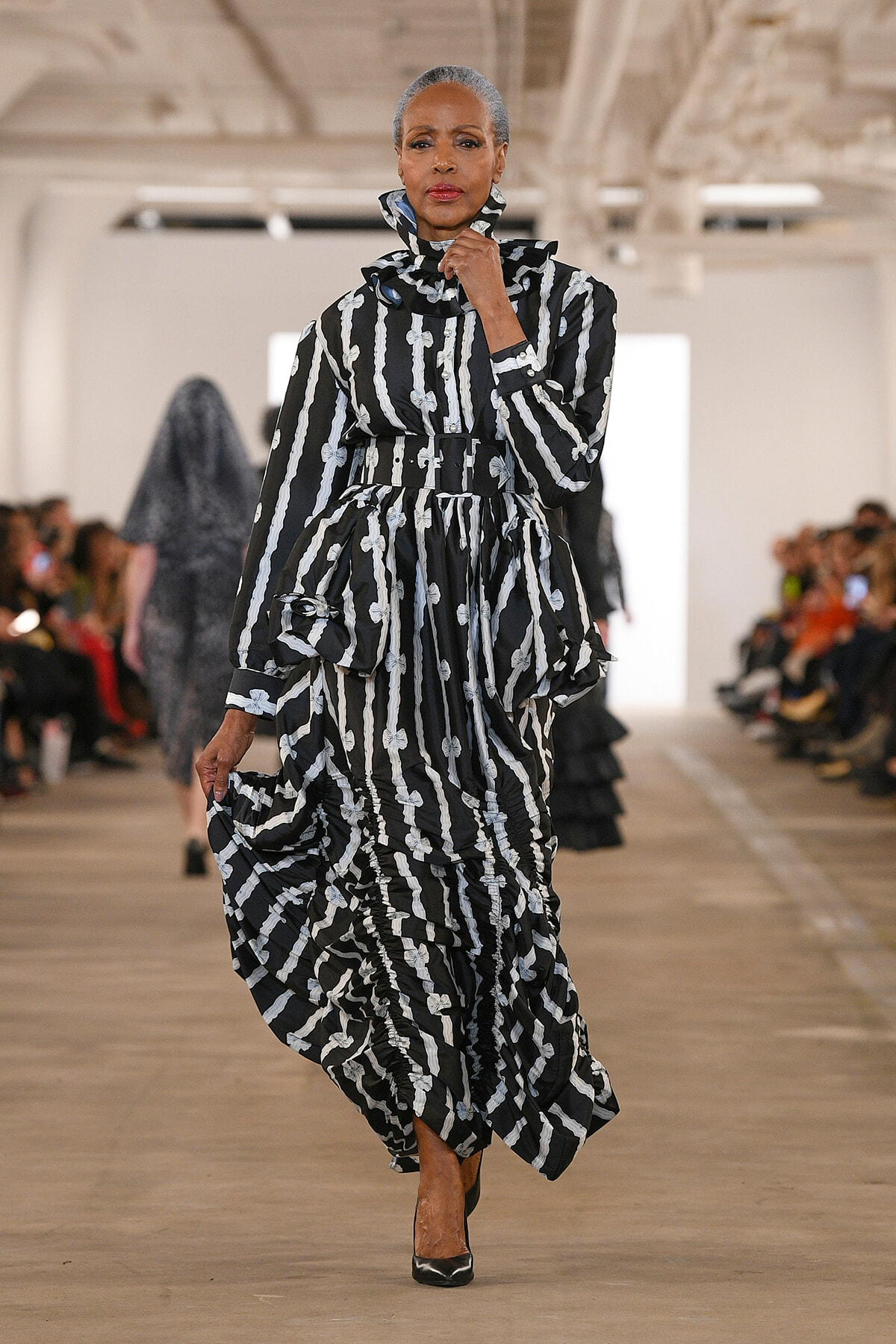 Model walks the runway in a black-and-white striped, ruffled dress with a high collar and belt; audience blurred in the background.