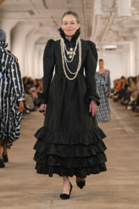 Older model on a runway wearing a black Victorian-inspired dress with layered ruffles and multiple pearl necklaces.