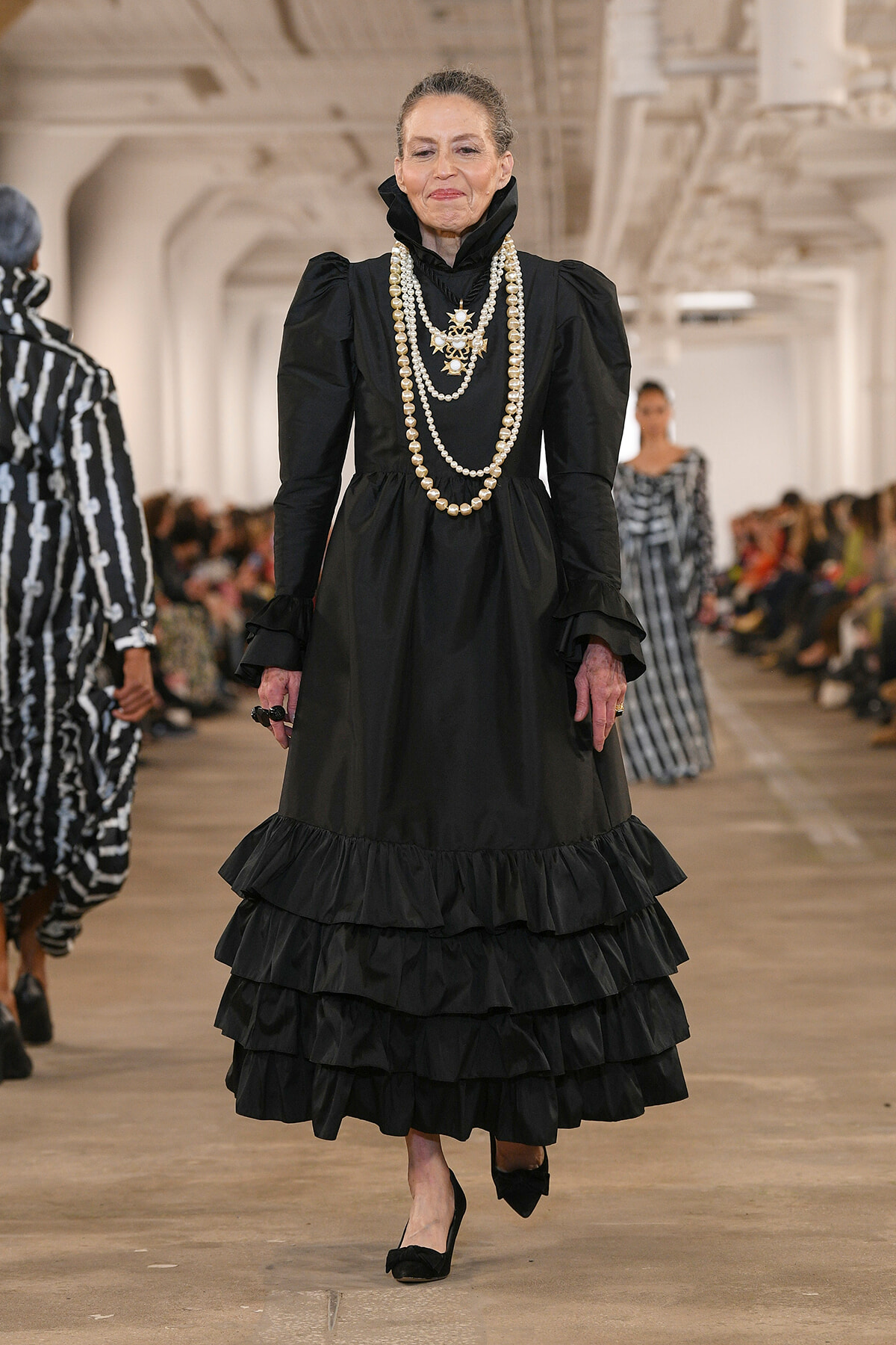 Older model on a runway wearing a black Victorian-inspired dress with layered ruffles and multiple pearl necklaces.