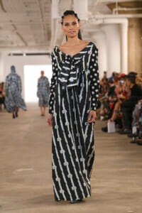 Model walking a runway in a black and white striped maxi dress with bow motifs, audience visible on the sides.