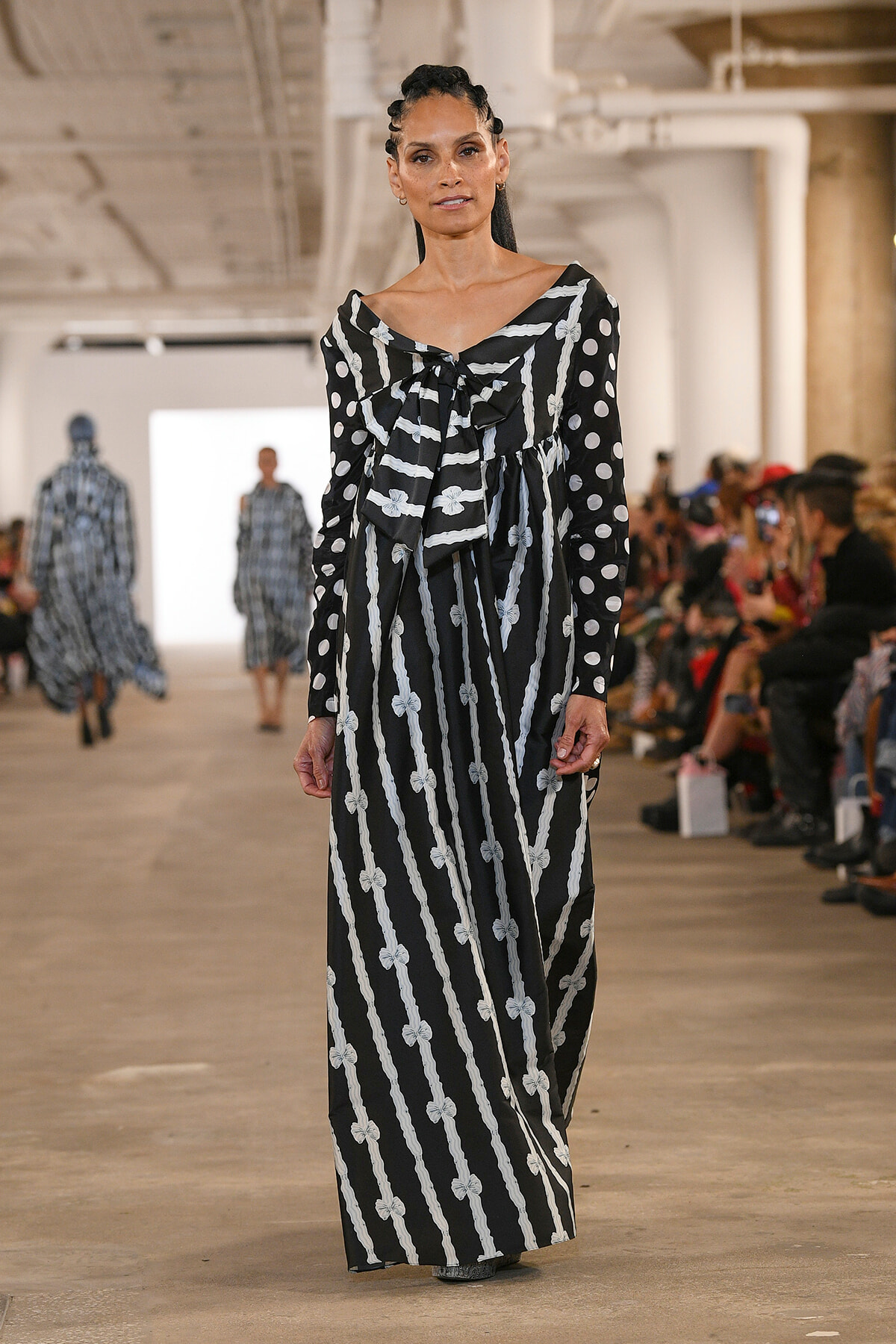 Model walking a runway in a black and white striped maxi dress with bow motifs, audience visible on the sides.