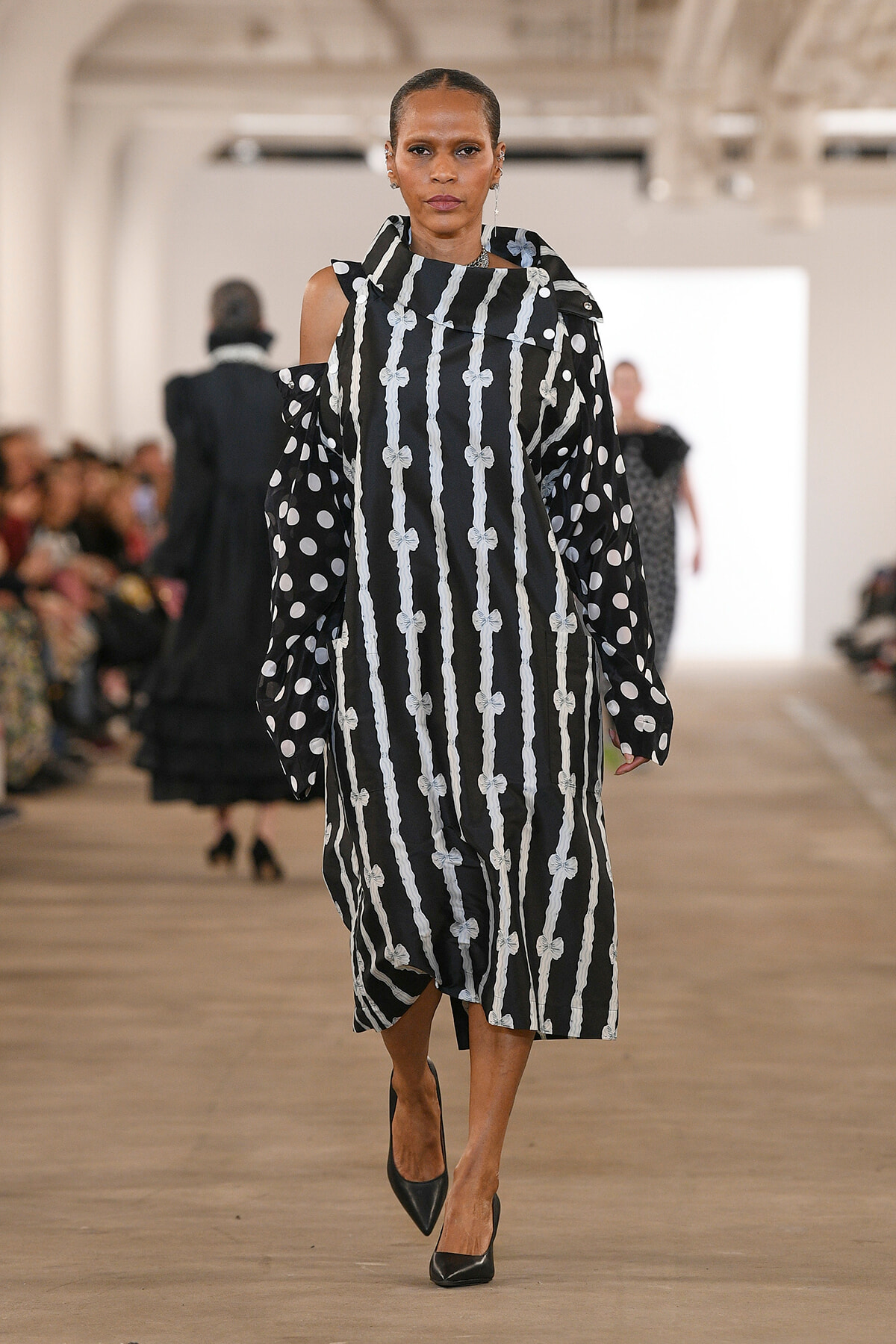 Model walking the runway in a black-and-white striped dress with bow-like accents and polka-dot sleeves.