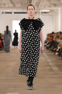 Model walking a runway in a black polka-dot dress with a large bow at the neckline.