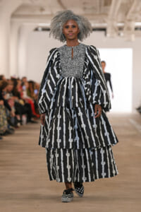 Model walks a fashion runway wearing a black-and-white striped, tiered dress with voluminous sleeves and a leopard-print bodice; blurred audience in background.