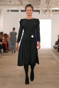 Model walking a runway in a long black dress with a white belt button, black tights and boots, audience in the background.