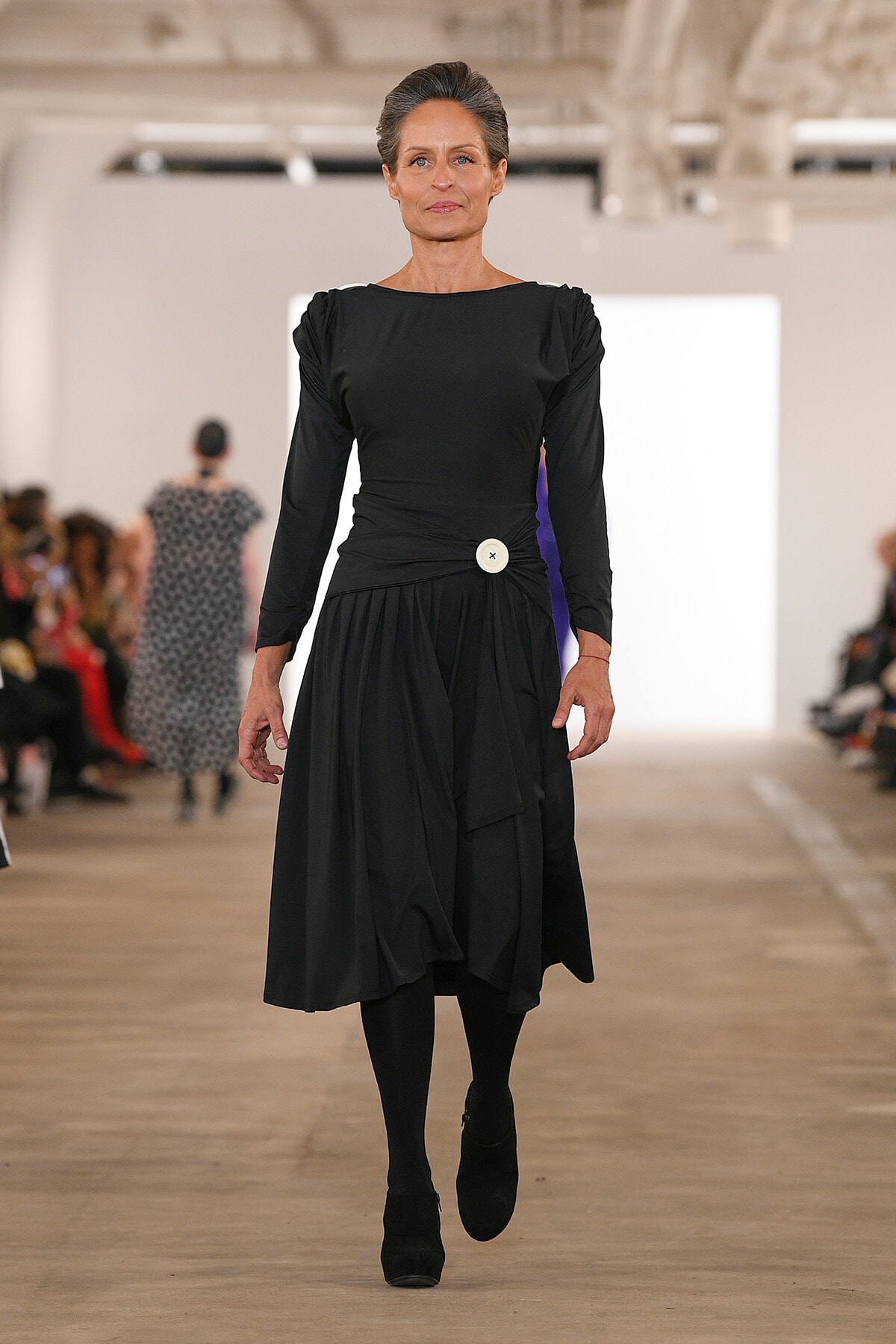Model walking a runway in a long black dress with a white belt button, black tights and boots, audience in the background.
