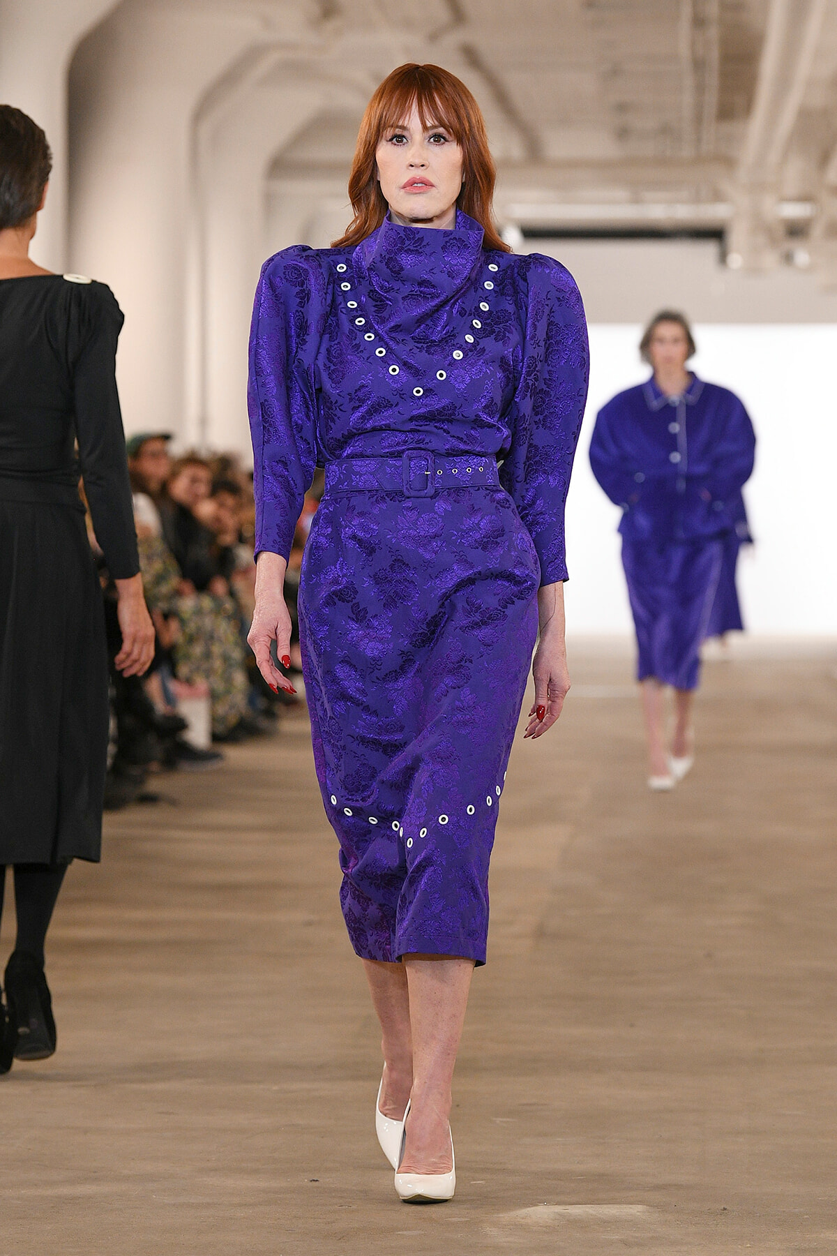 Runway model in a purple patterned dress with a belt and white heels walking toward the camera.
