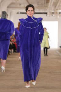 Model walks the runway in a royal blue velvet gown with white piping and ruffled collar, white heels visible.