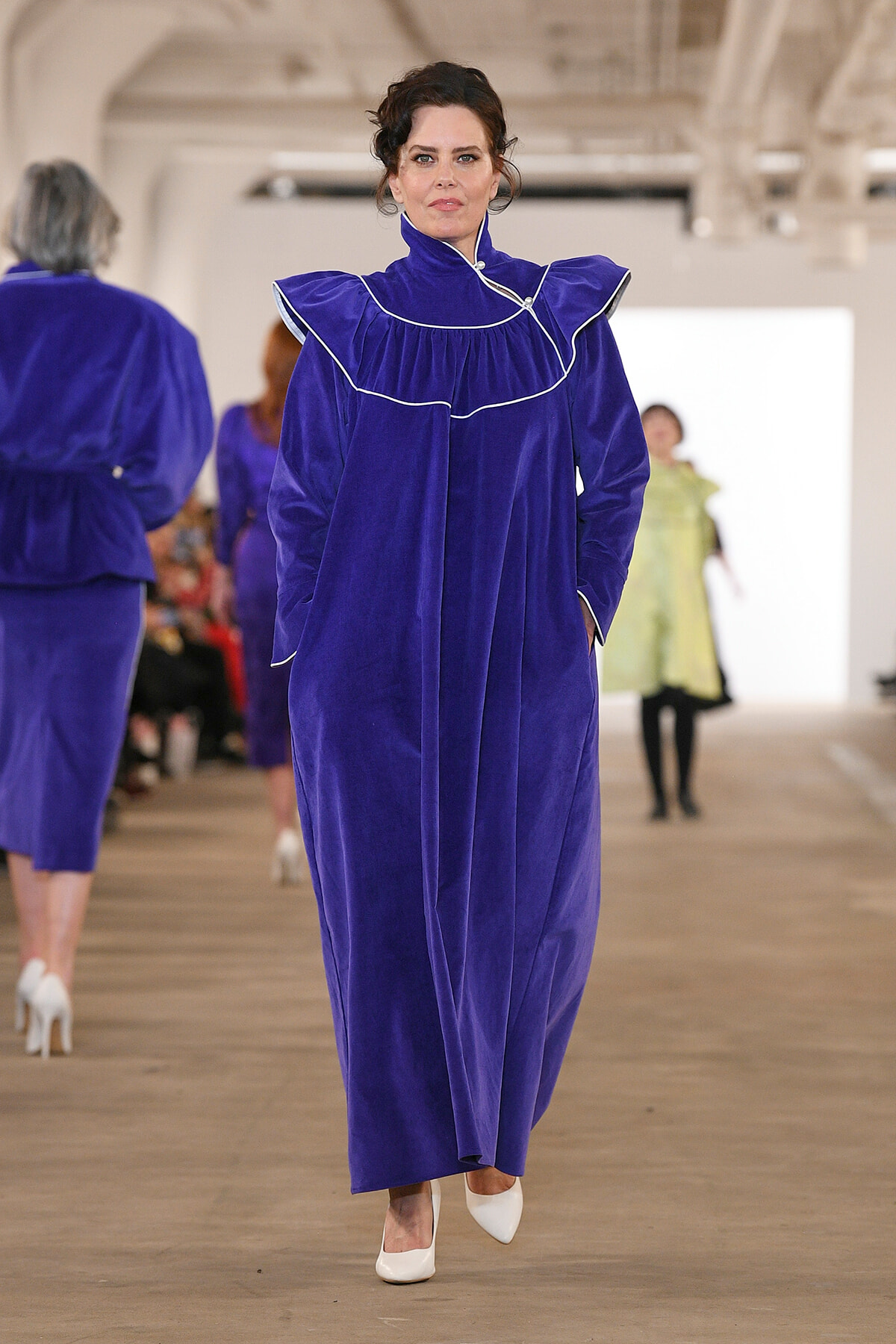 Model walks the runway in a royal blue velvet gown with white piping and ruffled collar, white heels visible.
