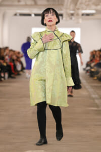Model walking a fashion runway in a pale green jacquard dress with black piping, hand resting on her chest as she strides forward.