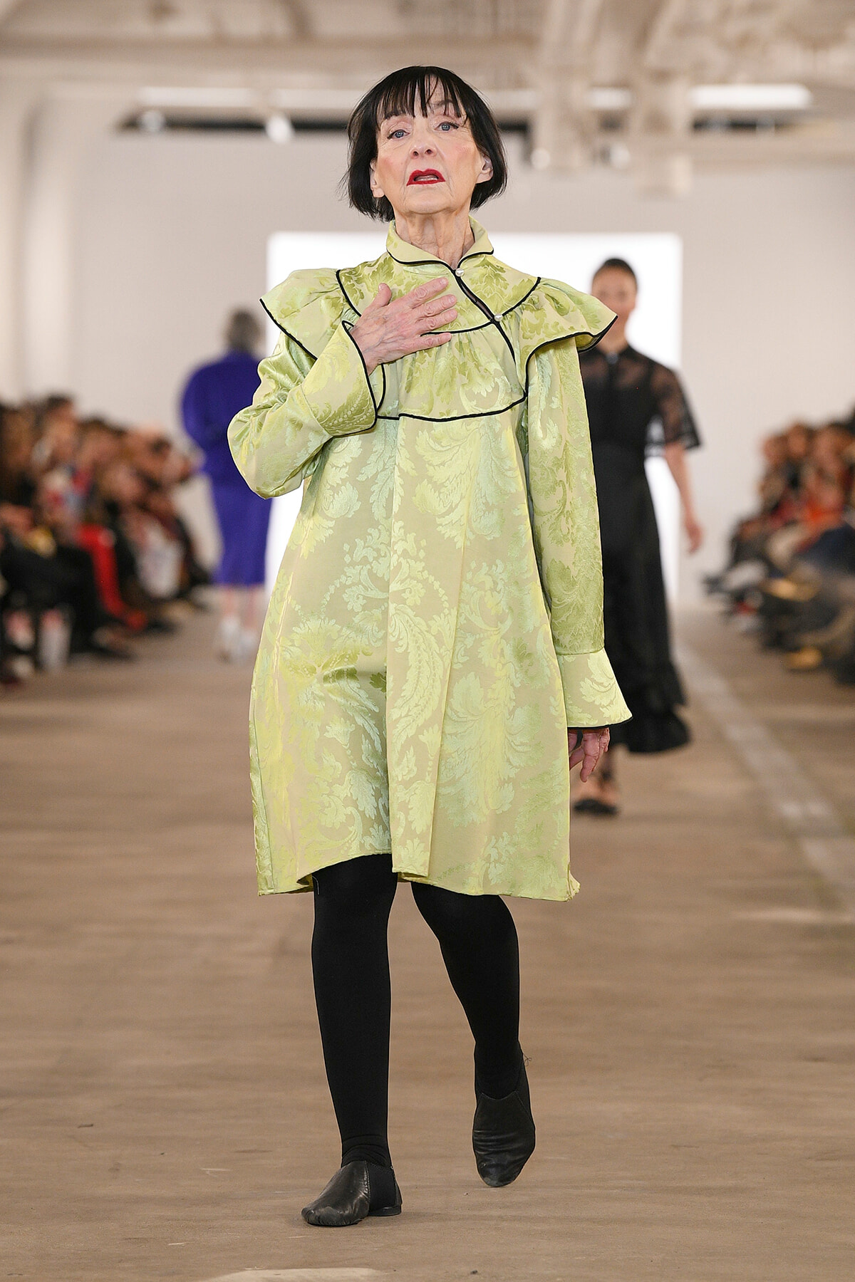 Model walking a fashion runway in a pale green jacquard dress with black piping, hand resting on her chest as she strides forward.