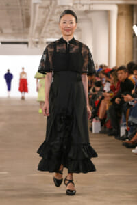 Female model walks the runway in a floor-length black gown with a sheer lace cape, audience on either side.