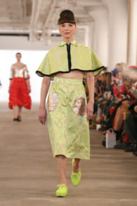 Model walks the runway in a lime-green damask-print cape-top and matching skirt with large sequin patches, paired with neon lime sneakers.