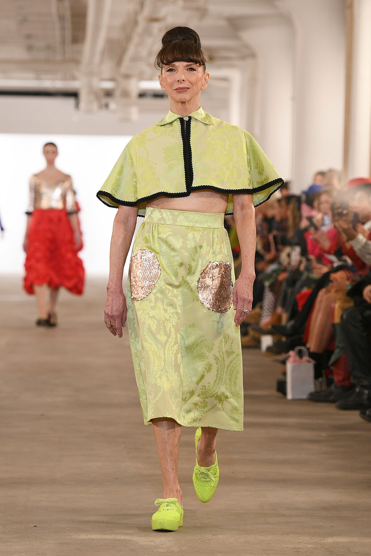 Model walks the runway in a lime-green damask-print cape-top and matching skirt with large sequin patches, paired with neon lime sneakers.