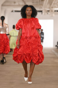 Female model walks a runway in a bright red oversized ruched dress with puff sleeves and white heels.
