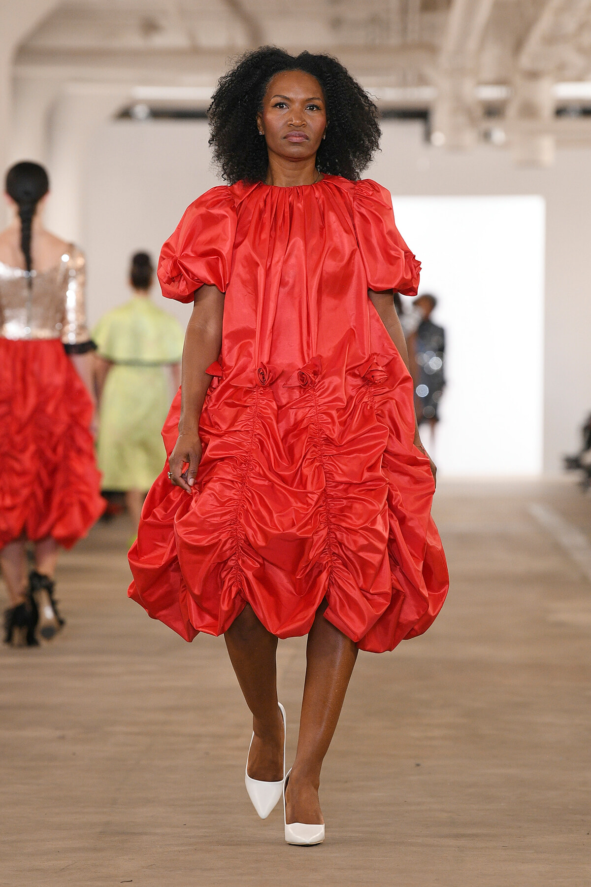 Female model walks a runway in a bright red oversized ruched dress with puff sleeves and white heels.