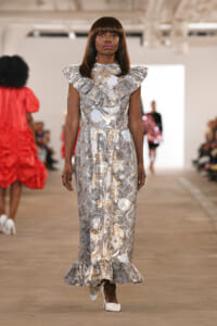 Model walks the runway in a silver-gray floral gown with ruffled shoulders and long skirt on a fashion show catwalk.