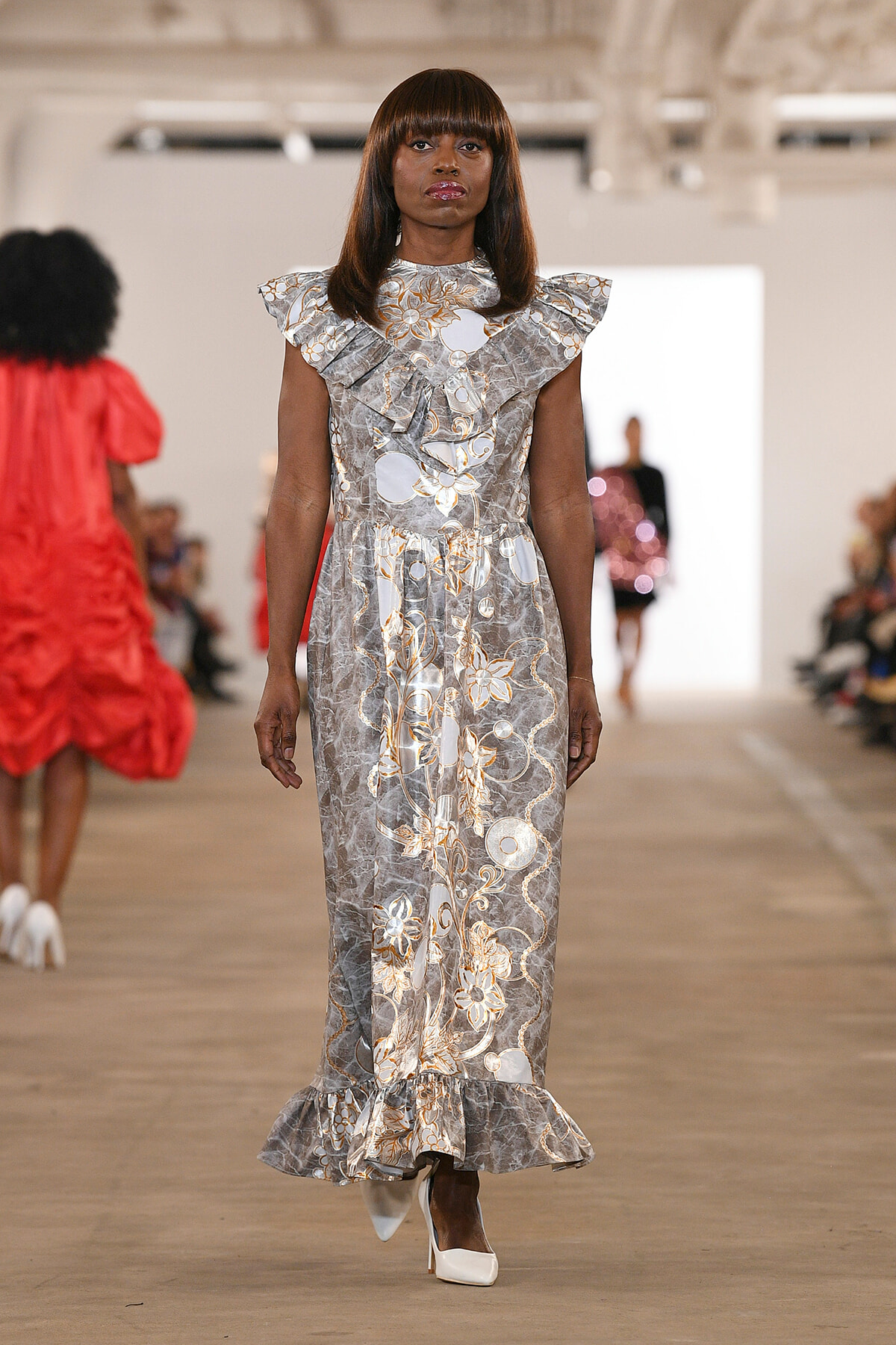 Model walks the runway in a silver-gray floral gown with ruffled shoulders and long skirt on a fashion show catwalk.