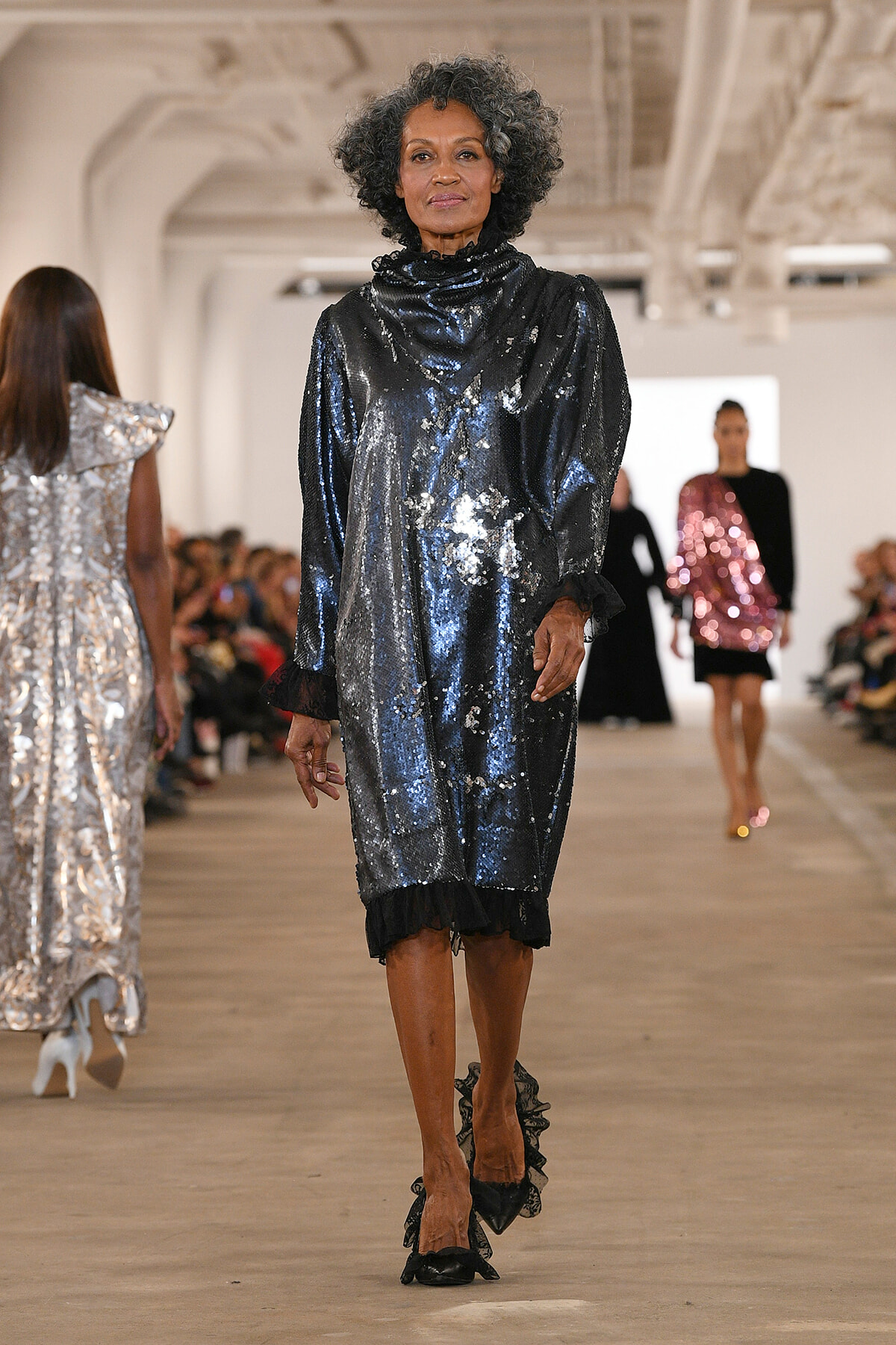 Model walks a fashion runway in a shiny blue-silver sequin dress with ruffled black cuffs and matching shoes, audience blurred in the background.
