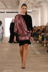 Model walking a runway in a pink sequin draped one-shoulder top over a black dress.