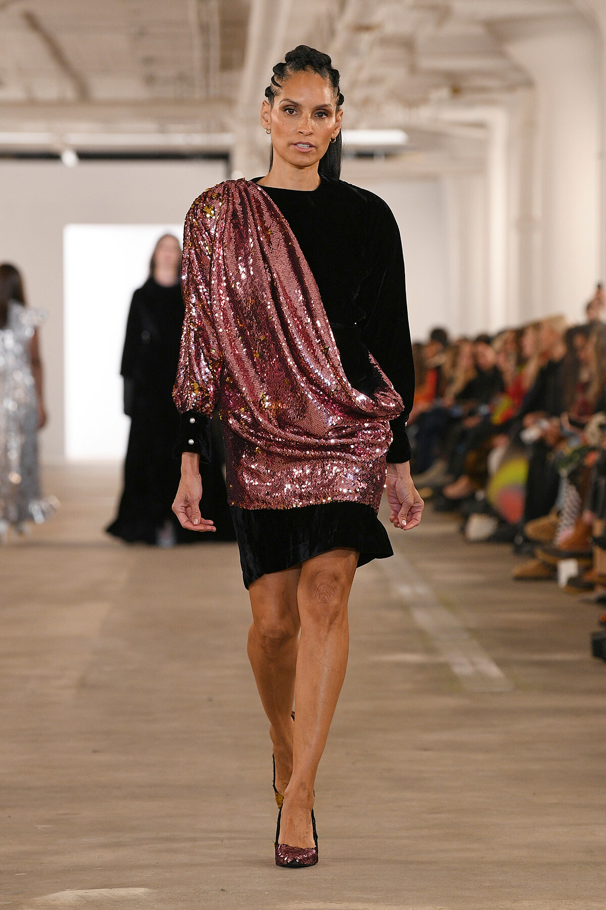 Model walking a runway in a pink sequin draped one-shoulder top over a black dress.