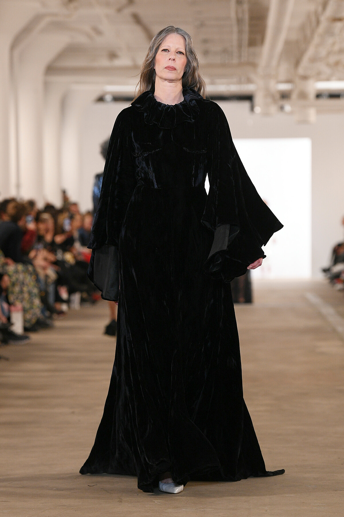 Model walks a runway in a floor-length black velvet gown with a ruffled neckline and dramatic bell sleeves.