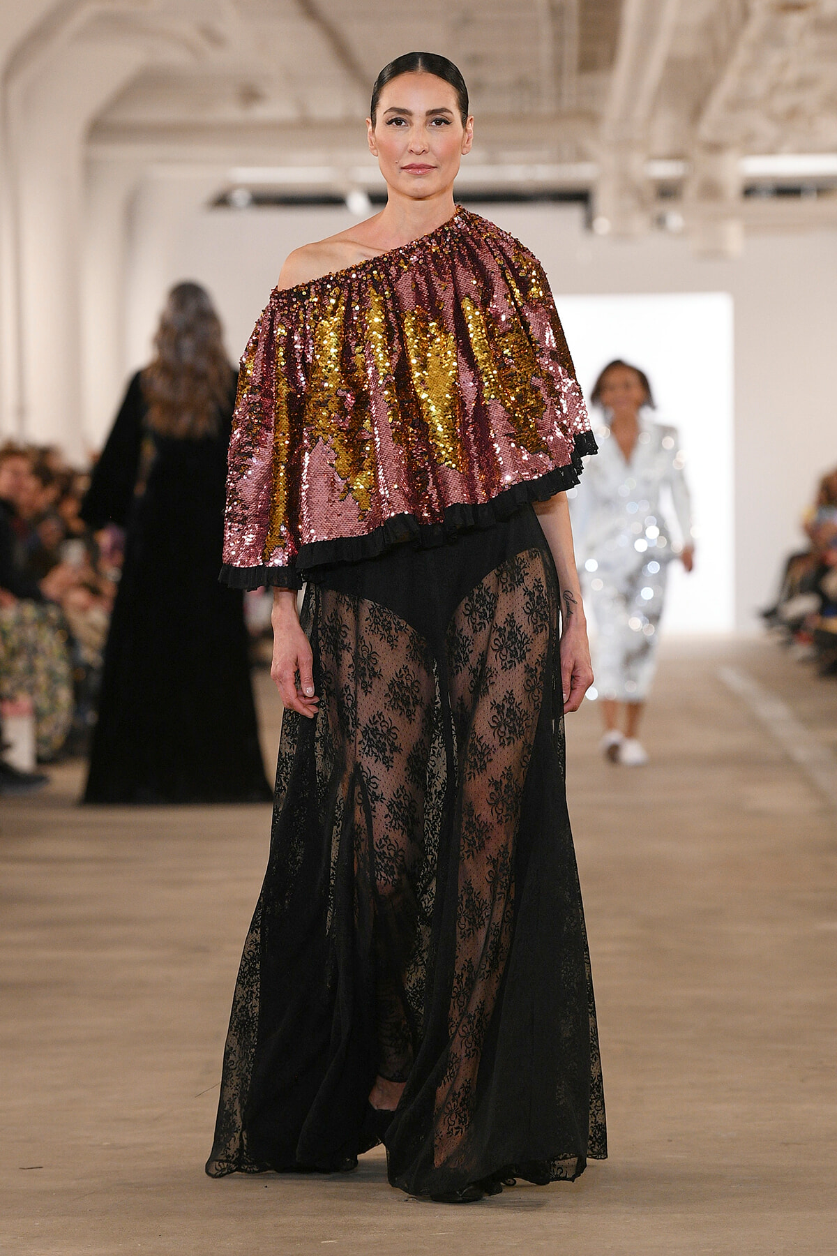 Model walking the runway in a burgundy-gold sequin one-shoulder top with a sheer black lace skirt.